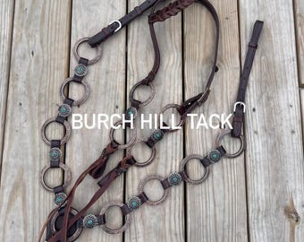 May include: A brown leather horse bridle with silver metal rings and turquoise accents. The bridle has decorative elements and the text "BURCH HILL TACK". The leather is a dark brown colour.