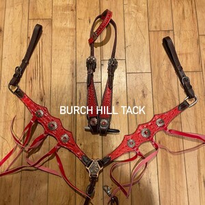 May include: Red and brown horse tack set with silver-toned accents. The set includes a headstall, breast collar, and reins. The breast collar and headstall feature decorative silver conchos. The text "BURCH HILL TACK" is visible.