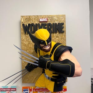 Op de afbeelding: Een driedimensionale wandbuste van het Marvel-personage Wolverine, met een geel en zwart pak, een masker met puntige oren en drie lange zilveren klauwen aan de rechterhand. De achtergrond is een goudkleurig, getextureerd paneel.