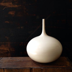 May include: A white ceramic vase with a rounded, bulbous body and a long, thin neck. The vase is sitting on a wooden surface.
