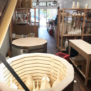 May include: Interior shot of a pottery studio with a kiln in the foreground and shelves filled with white ceramic vases and sculptures. The studio has a bright, open feel with natural light and a view of the outside.