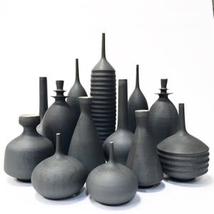 May include: A collection of dark gray ceramic vases of varying shapes and sizes. Some vases have a smooth finish, while others feature textured or ribbed designs. The vases are arranged against a white background, showcasing their unique forms.