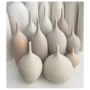 May include: A collection of ceramic vases in various neutral tones, including beige, cream, and off-white. The vases have a minimalist design with elongated necks and rounded bodies. Some vases are tall and slender, while others are more bulbous.