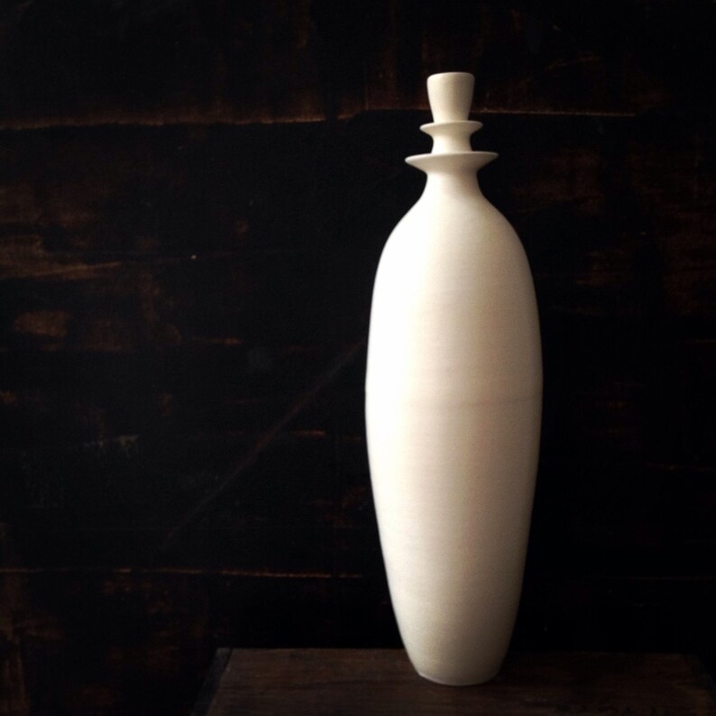 May include: A tall, off-white ceramic bottle with a decorative stopper. The bottle has a rounded body and a long, slender neck. The stopper is composed of stacked, circular elements. The background is a dark, textured surface.