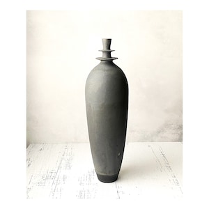 May include: A tall, gray ceramic vase with a unique, layered top. The vase has a smooth, slightly textured surface and a dark gray base.