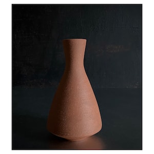 May include: A terracotta-colored ceramic vase with a wide base and a narrow neck. The vase has a rough, textured surface.