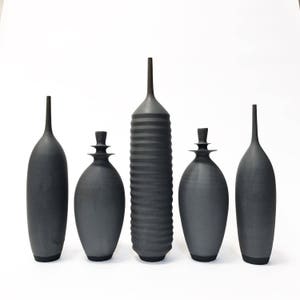 May include: Five black ceramic vases of varying shapes and sizes. The vases are arranged in a row on a white surface.