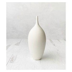 May include: A white ceramic vase with a rounded, bulbous body and a long, slender neck. The vase is sitting on a white wooden surface.