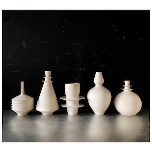 May include: Five white ceramic vases of varying shapes and sizes are arranged in a row. The vases have a matte finish and are set against a dark background. The designs range from a simple bottle shape to more complex, tiered forms.