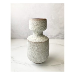 May include: A textured, off-white ceramic vase with a unique hourglass shape. The vase has a wide base, a narrower midsection, and a flared top. The rim and the base of the top section are a darker, earthy brown color. The surface has a speckled, porous texture.
