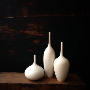 May include: Three white ceramic vases of varying shapes and sizes. The vases are arranged on a wooden surface against a dark background.