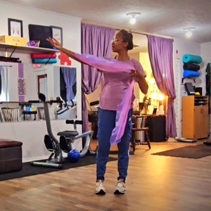May include: A woman wearing a pink shirt and blue pants is holding a purple scarf and looking to the right. She is standing in a room with a wooden floor and a mirror in the background.