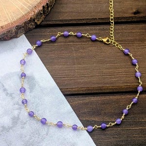 May include: A delicate gold-tone chain anklet featuring small, round, light purple amethyst beads spaced evenly along the wire. The anklet includes a gold-tone extender chain with a lobster claw clasp, creating a circular shape on a dark surface.