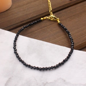 May include: A delicate bracelet featuring small, faceted black beads strung together. The piece includes a gold-toned metal lobster claw clasp and an adjustable chain link extension, providing a secure and customizable fit for the wearer's wrist.