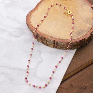 May include: A delicate gold chain necklace featuring small, round, ruby-colored beads. The necklace is displayed on a wooden surface with a marble background, showcasing the jewelry's design and color.