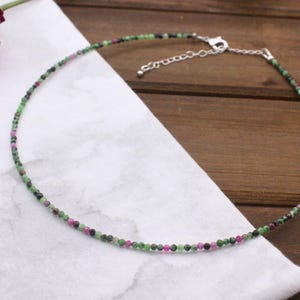 May include: Delicate beaded necklace featuring small, round gemstones in shades of green, pink, and black. The necklace has a silver-colored clasp and is displayed on a white cloth against a wooden background. A simple and elegant piece of jewelry.
