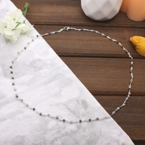 Può includere: Una collana delicata caratterizzata da una sottile catena color argento intervallata da piccole perline rotonde di agata muschiata verde scuro. La collana è fissata con un piccolo moschettone in metallo, creando un design circolare.