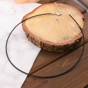 Könnte beinhalten: Eine zarte Halskette aus schwarzen Perlen mit einem silbernen Verschluss, präsentiert auf einer Holzoberfläche. Die Halskette besteht aus kleinen, dunklen Perlen, die ein Choker-Design bilden. Der Hintergrund umfasst eine Holzscheibe und ein weißes Tuch.