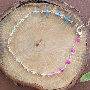 May include: A delicate anklet with a rose gold chain and colorful glass beads. The beads are arranged in a gradient pattern, starting with yellow, transitioning to blue, and ending with pink.