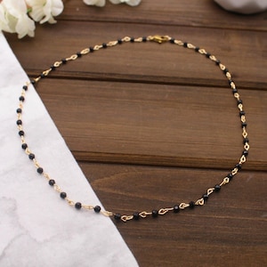 May include: A delicate gold-tone necklace featuring small black beads strung along a fine chain. The necklace is displayed on a wooden surface with a white cloth and a few white flowers in the background. The necklace has a clasp closure.