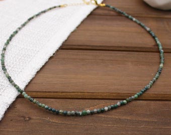 May include: A delicate necklace composed of small, round, variegated green and translucent moss agate beads. The strand features a gold-toned metal lobster claw clasp and a short extender chain, creating a simple, circular beaded design.
