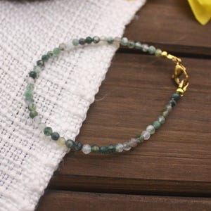 May include: Delicate beaded bracelet featuring small, faceted green and clear gemstones. The bracelet has a gold-colored clasp and is displayed on a white cloth and a wooden surface. The bracelet is a delicate piece of jewelry.