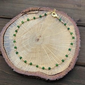 May include: A gold chain anklet with green beads. The anklet is on a wooden surface.