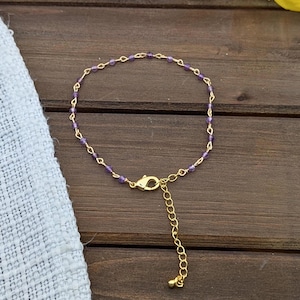 May include: A delicate gold-tone chain bracelet featuring small, round, purple amethyst beads spaced evenly along the links. The bracelet includes a gold-tone lobster claw clasp and an adjustable extender chain with a small bead at the end.