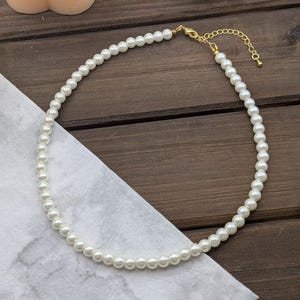 May include: A white pearl necklace with a gold clasp and extender chain. The necklace is on a wooden surface.