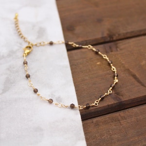 May include: A delicate gold-toned chain bracelet featuring small, faceted dark brown beads spaced evenly along the links. The bracelet includes a lobster claw clasp and an adjustable extender chain with a small dangling charm at the end.