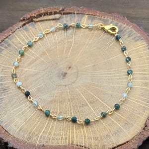 May include: A delicate gold bracelet with small, round beads in shades of green and clear. The beads are strung on a fine gold chain, secured with a gold clasp. The bracelet is displayed on a wooden surface. The bracelet is approximately 18 cm in length.