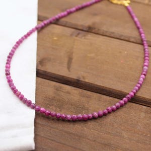 May include: A delicate necklace composed of small, faceted magenta-colored beads arranged in a single strand. The necklace features a gold-toned metal clasp at the end, providing a subtle contrast to the vibrant, textured surface of the circular beads.