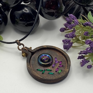 May include: A handcrafted wooden pendant necklace featuring a circular design with a central iridescent button. The pendant has embroidered details in green and purple thread. The necklace has a black cord and a copper-colored clasp.