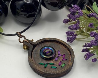 May include: A handcrafted wooden pendant necklace featuring a circular design with a central iridescent button. The pendant has embroidered details in green and purple thread. The necklace has a black cord and a copper-colored clasp.