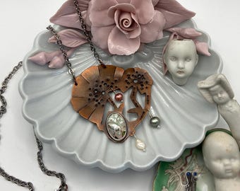 May include: A copper-colored pendant necklace with a heart-shaped design, featuring a silver chain and a pear-shaped pearl. The necklace rests on a gray ceramic dish with a pink floral accent and doll heads.