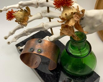 May include: A copper cuff bracelet with three green gemstones, resting on a dark gray block. The bracelet has a textured surface and is next to a small green glass bottle and a decorative skeleton hand with dried flowers.
