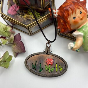 May include: A wooden pendant necklace with an oval embroidery hoop design. The pendant features a copper-toned background with embroidered green stems and a red flower. The necklace has a black cord and a copper-colored clasp.