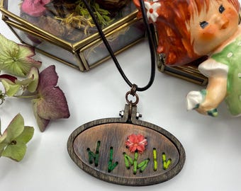 May include: A wooden pendant necklace with an oval embroidery hoop design. The pendant features a copper-toned background with embroidered green stems and a red flower. The necklace has a black cord and a copper-colored clasp.