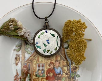 May include: A circular pendant featuring a dark wood embroidery hoop frame containing white fabric with green and blue stitched details. The pendant hangs from a black cord and is positioned over a white plate decorated with a colorful illustration.