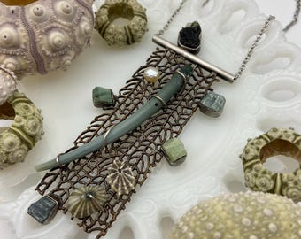 May include: A necklace featuring a rectangular, open-work bronze metal mesh pendant adorned with a central green tube, a small white pearl, and various small green mineral stones. The pendant is suspended from a thin silver-toned metal chain.