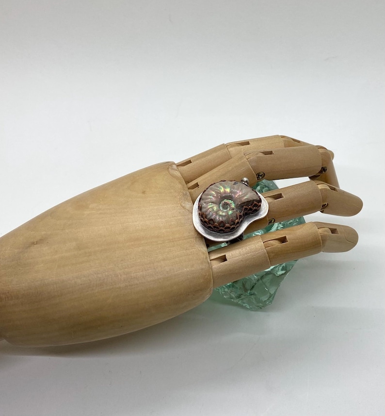 May include: A silver ring with a copper and iridescent ammonite gemstone. The ring is set on a wooden hand model.