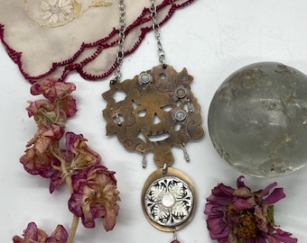 May include: A silver chain necklace with a copper pendant featuring a skull design with floral accents and a pearl-like button. The pendant is attached to a silver chain with a pearl-like bead.