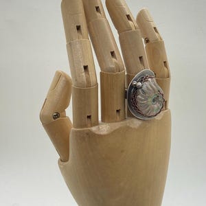 May include: A silver ring with a large, iridescent, pink and white shell-like design. The ring is set on a wooden hand model.