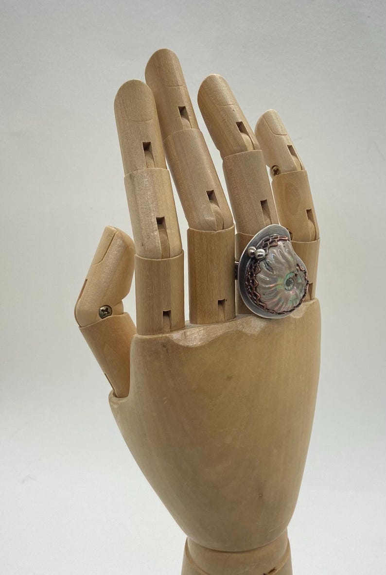 May include: A silver ring with a large, iridescent, pink and white shell-like design. The ring is set on a wooden hand model.