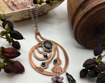 May include: A copper-colored pendant necklace with an oval, dark gray stone set in a silver bezel. The pendant has an openwork design with black and purple accents, and a chain. The necklace is displayed with dried berries and a decorative wooden object.