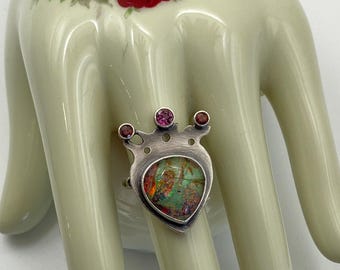 May include: A silver ring with a heart-shaped, multi-colored stone. The ring has a crown design with three small, round gemstones. The ring is displayed on a cream-colored hand with a floral design.