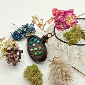 May include: A wooden oval pendant with a green, blue, and white embroidered design. The pendant is attached to a black cord and is surrounded by dried flowers and moss.