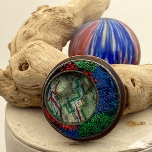 May include: A round copper-colored pendant with a map of Estes Park, Colorado inside. The map is surrounded by a border of colorful fibers in shades of red, green, and blue.