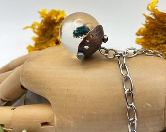 May include: A pendant featuring a clear spherical resin orb containing a small iridescent green beetle. The orb is set into a dark brown textured cap with metallic accents, attached to a silver-toned metal chain link necklace.