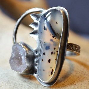 May include: A silver ring with a large, oval-shaped agate stone set in the center. The agate is a light gray color with black and white markings. A smaller, clear quartz stone is set on the side of the ring.